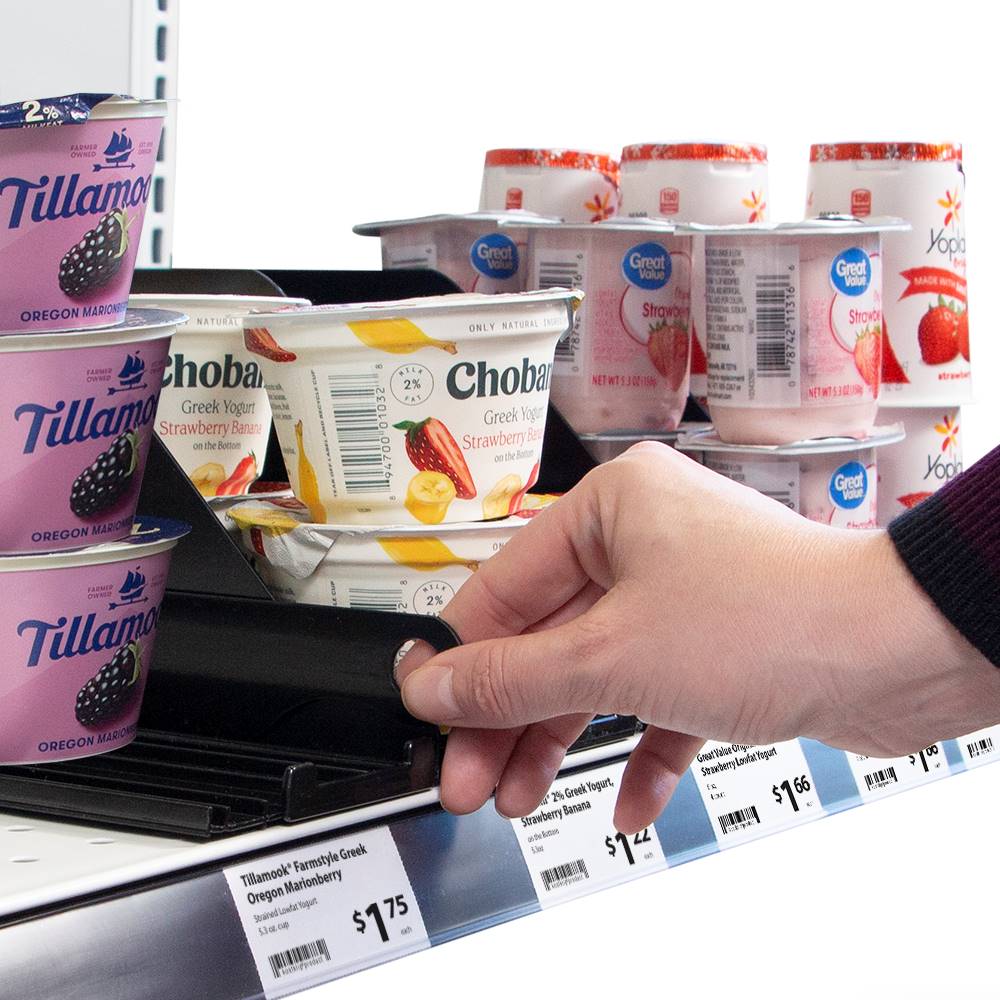The wide Yogurt Tender KwikPull System installed on a shelf holding different types of yogurt and a hand pulling the lever foward to front face the yogurt